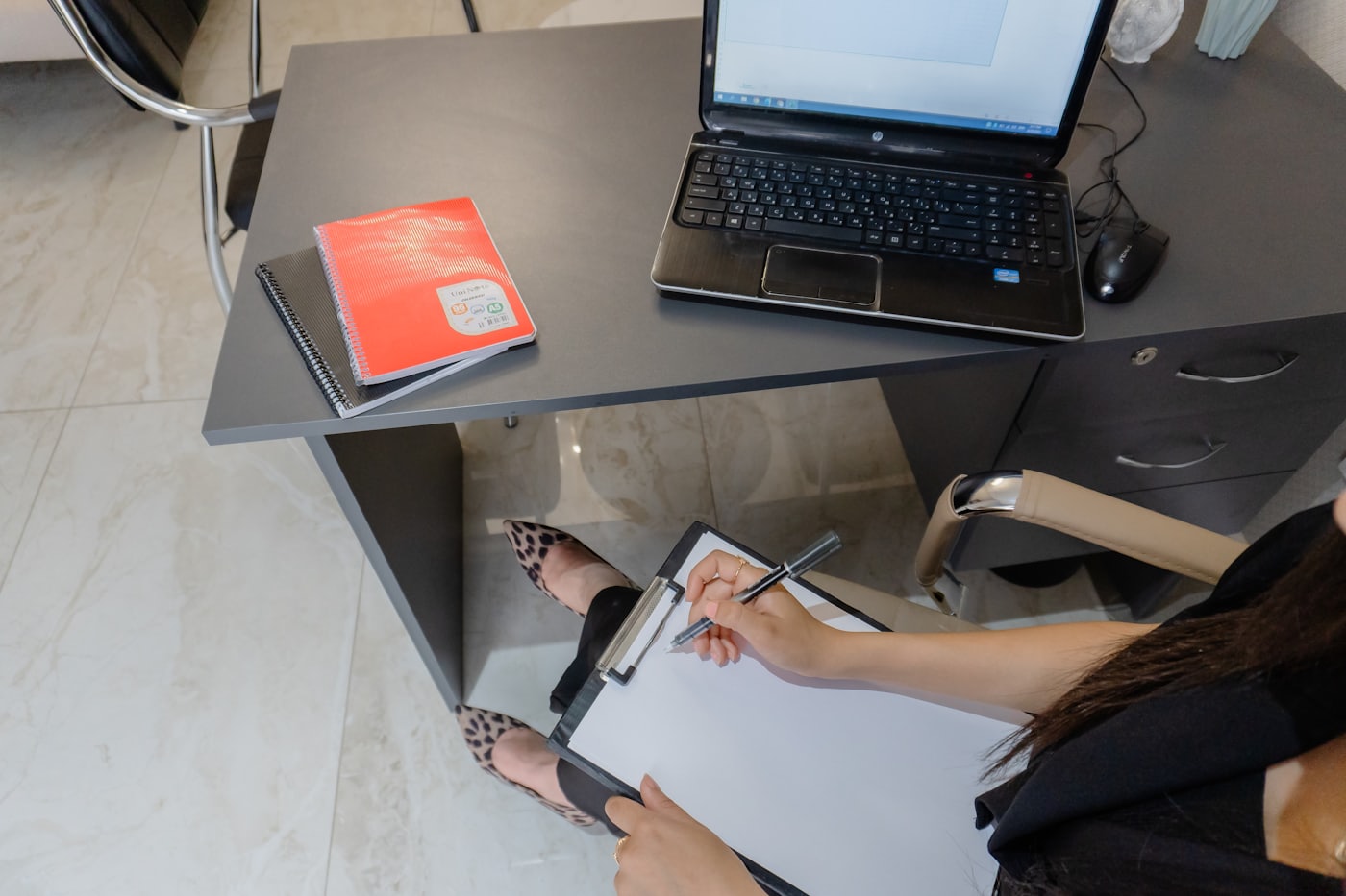 A person writing on a clipboard beside a laptop at a desk